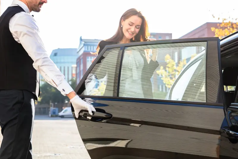 A professional chauffeur in uniform opens the door for an elegantly dressed, smiling young woman entering the back seat of a black luxury sedan, symbolizing Houston to Galveston luxury transportation.